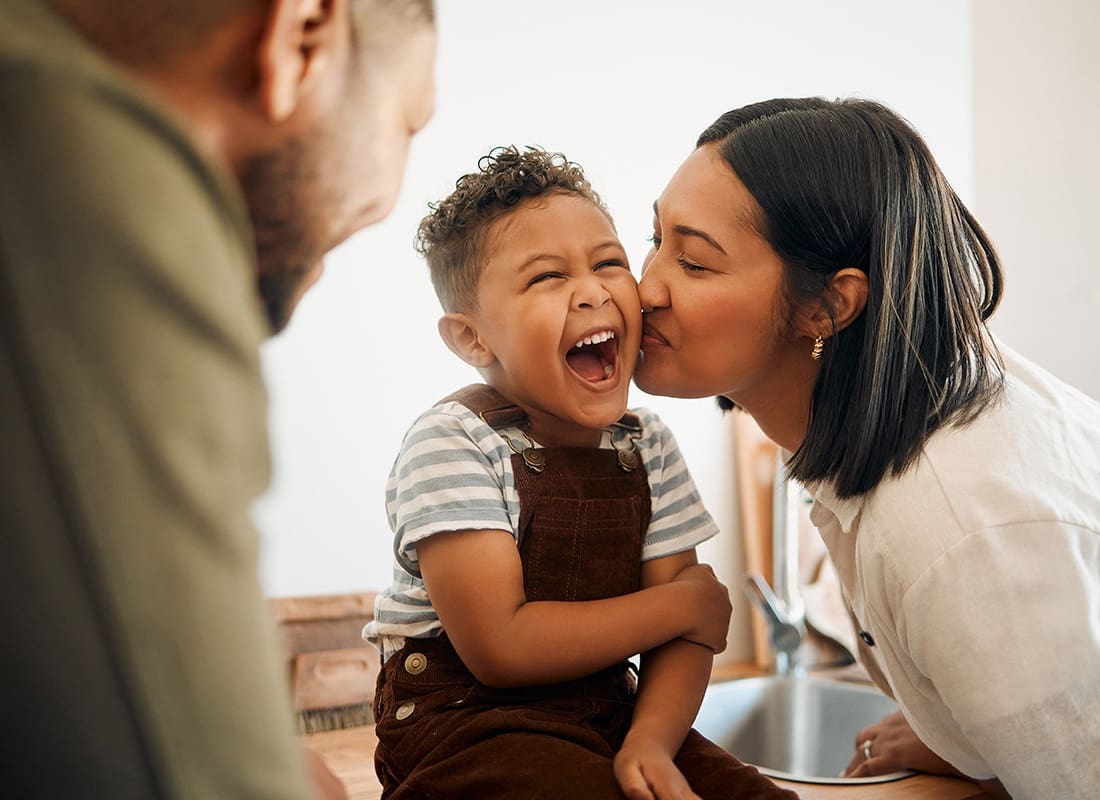 Contact - Portrait of a Mother Kissing her Excited Young Son Sitting on the Kitchen Counter on the Cheek Next to the Father